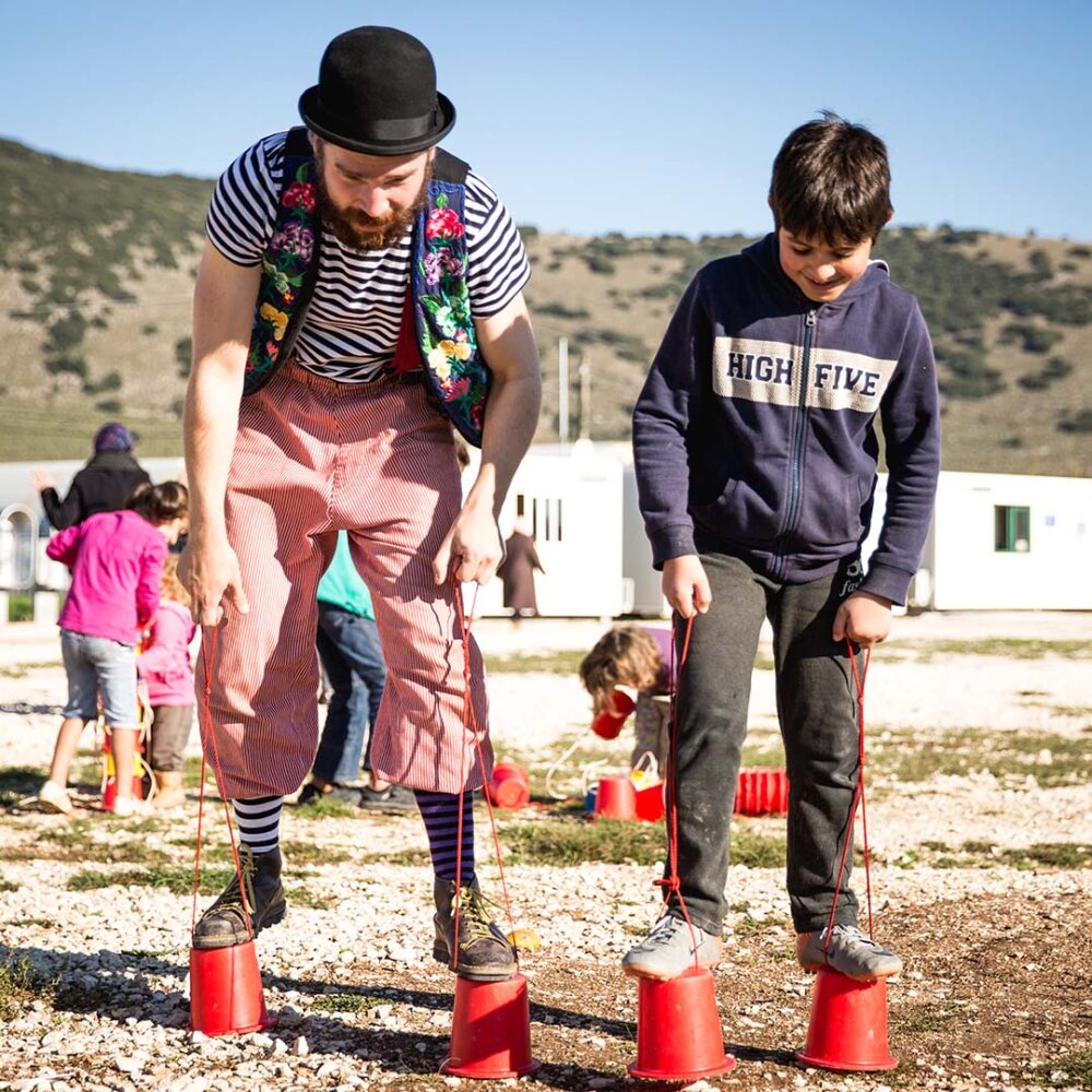 A child in a refugee camp smiles as he learns how to walk on stilts.